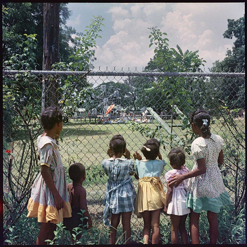 Five young Black girls and one infant in 1950's America looking through a metal fence at a playground in distance filled with White children and families playing.