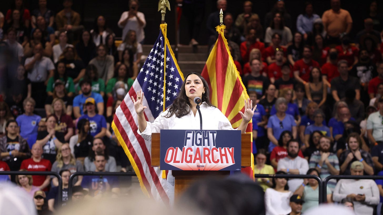 Rep. Alexandria Ocasio-Cortez speaking in front of a US flag and an Arizona flag and a packed stadium audience. She's at a podium with the words "Fight Oligarchy" on it. Rep. Alexandria Ocasio-Cortez speaking in front of a US flag and an Arizona flag and a packed stadium audience. She's at a podium with the words "Fight Oligarchy" on it.