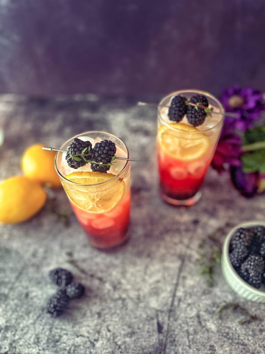 Two purple and gold mocktails in tall skinny glasses, garnished with lemon wheels and skewered blackberries