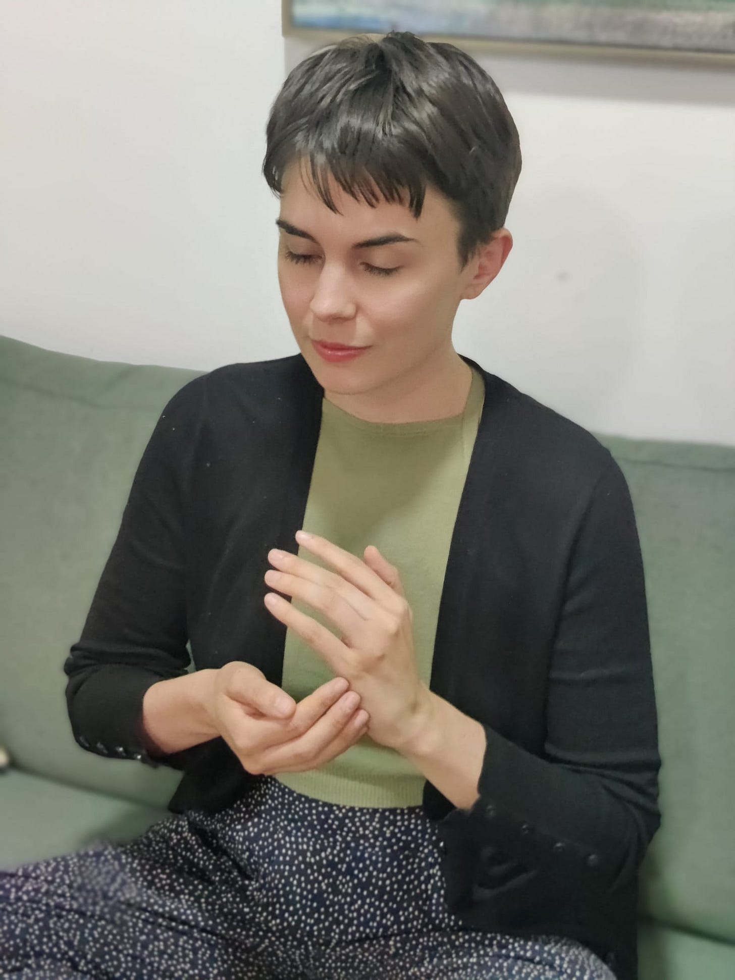 Caitlin has short brown hair and is wearing a green shirt. She sits on a dark green couch cross-legged with her eyes closed while tapping on the side of her hand.