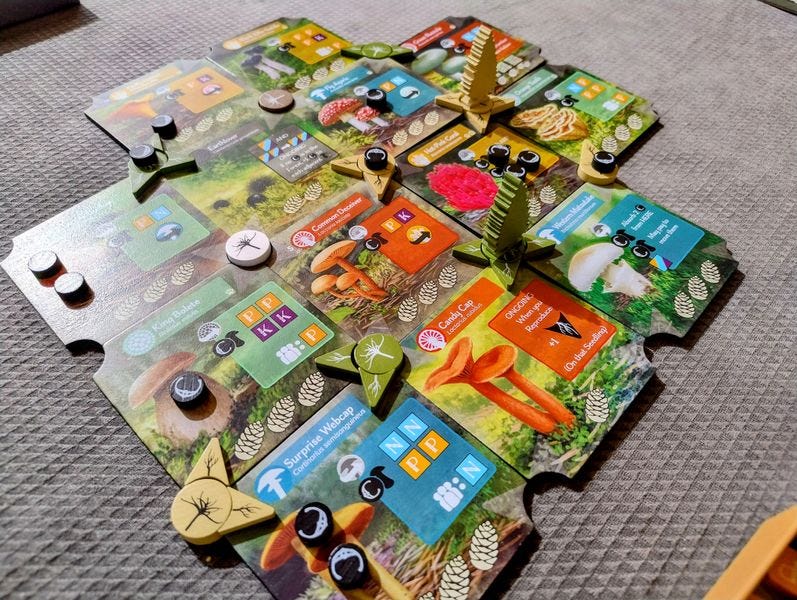 A game of Undergrove in progress. There are 13 mushroom tiles on the table, with a variety of player pieces and resources placed on them. 