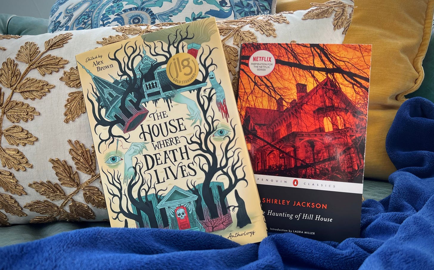 the books THE HOUSE WHERE DEATH LIVES edited by Alex Brown and THE HAUNTING OF HILL HOUSE by Shirley Jackson side by side against a stack of gold, blue, and teal pillows and blankets