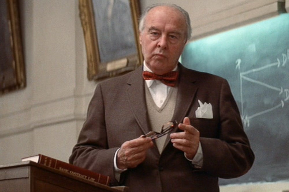 Screenshot of John Houseman in 'The Paper Chase,' standing at a lectern, holding his folded reading glasses, and looking sour