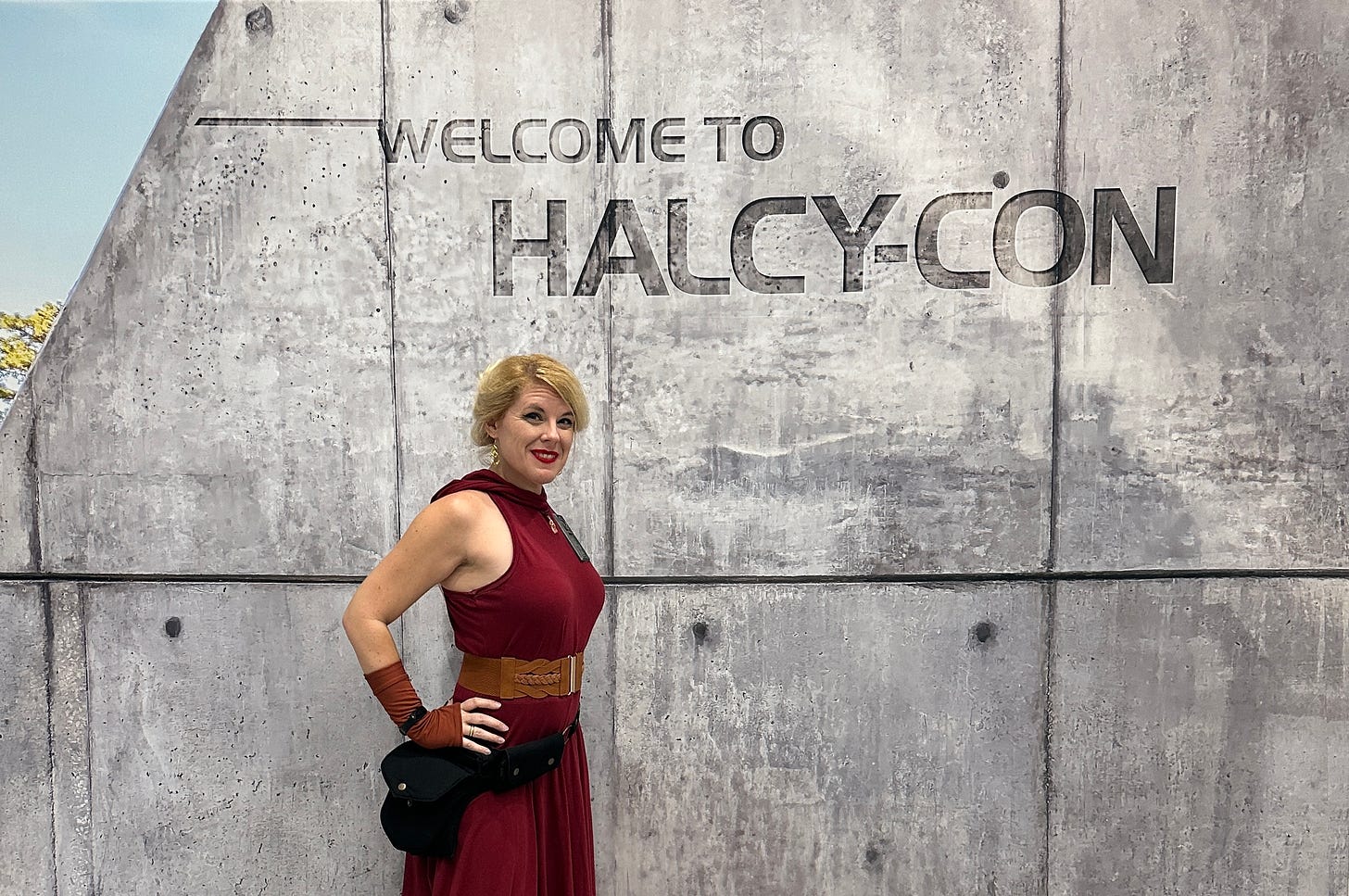 Cass in a red dress with Star Wars-y accessories standing in front of a grey industrial concrete-esque backdrop with the words "Welcome to Halcy-Con" on it