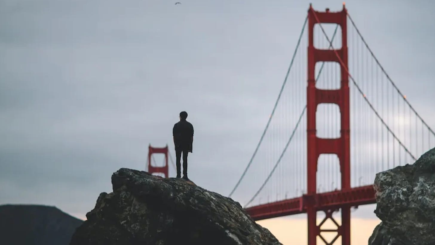 man stands on rock by golden gate bridge. you see him at a distance, back to the camera, looking over the water and silhouetted by the morning light