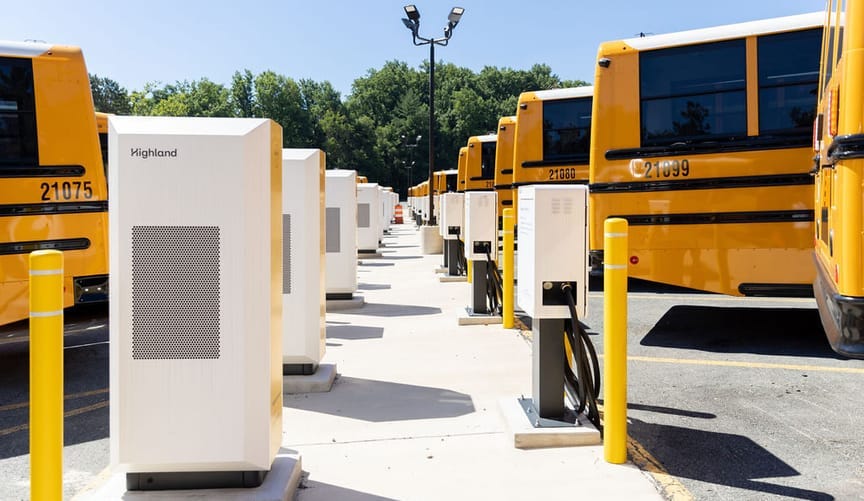 A parking lot full of many electric school buses, some of which are hooked up to chargers