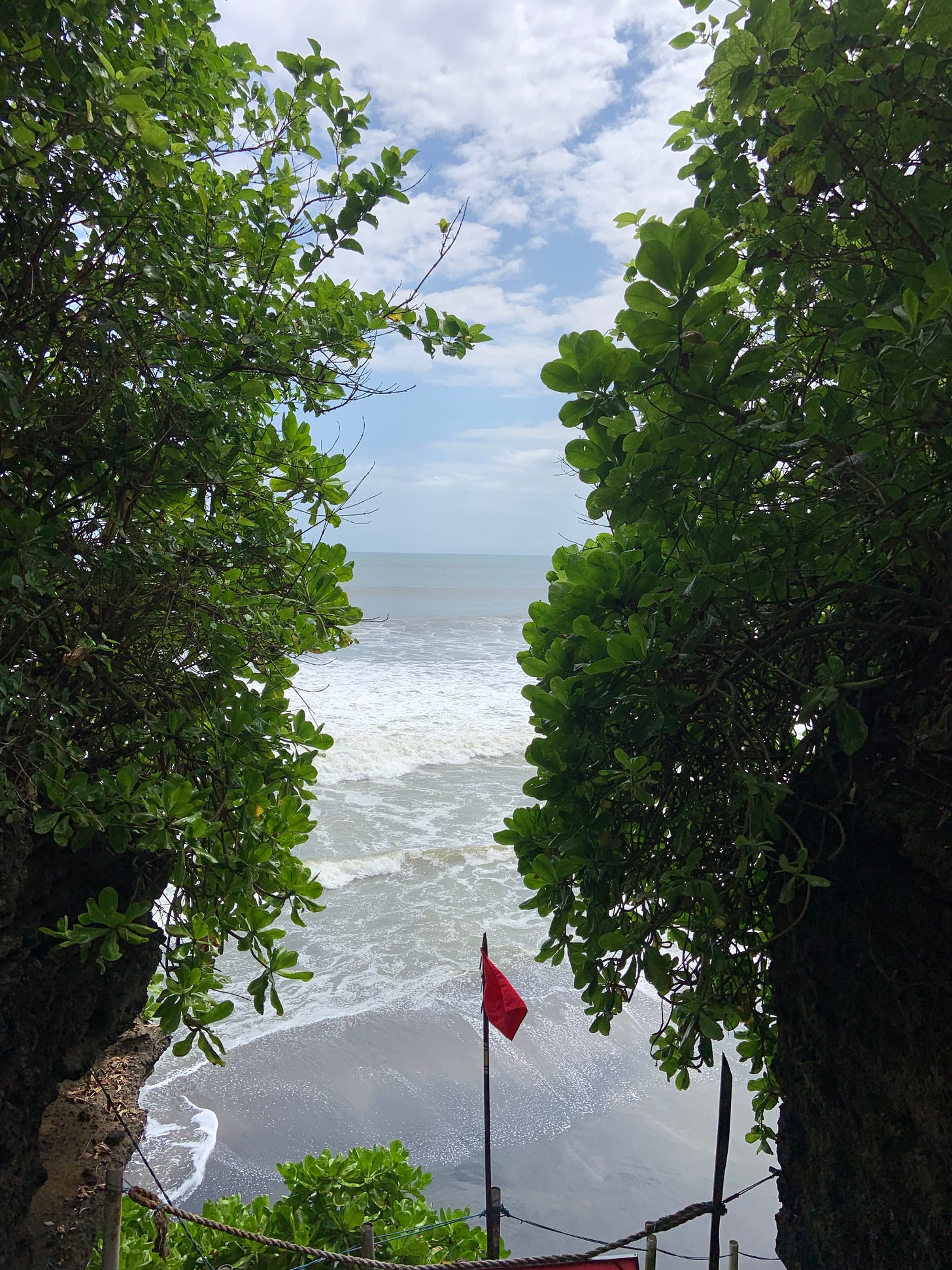 a photo of the beach captured in between two green trees; a red flag is posted in the middle signaling a high tide