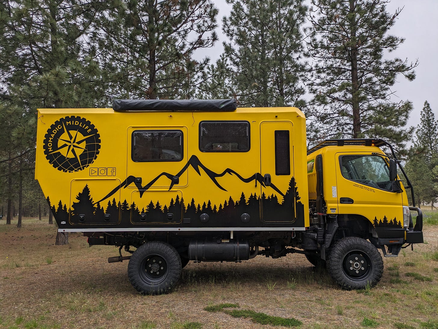 a big yellow camper truck built on a medium duty commercial chassis, decorated with black outlines of trees, mountain peaks, and a big logo that says Nomadic Midlife