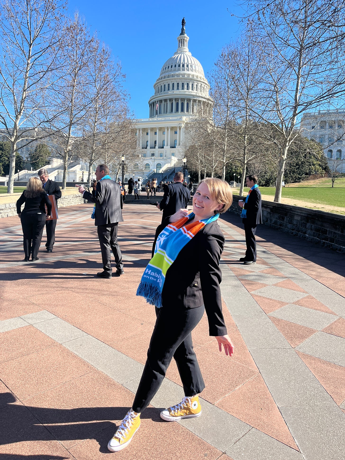 Christine is standing in front of the U.S. Capitol wearing a black suit, bright colored Habitat scarf and yellow high-top Converse.