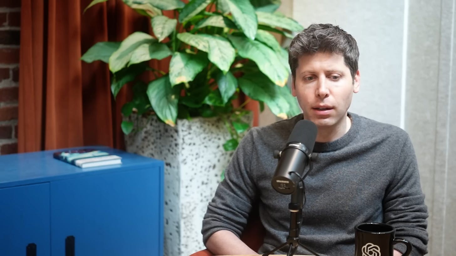 The image features Sam Altman sitting in an indoor setting, while he is recorded for the podcast. The individual appears to be speaking or pausing momentarily while speaking. They are wearing a grey sweater and are seated in front of a microphone with a pop filter attached to a stand.