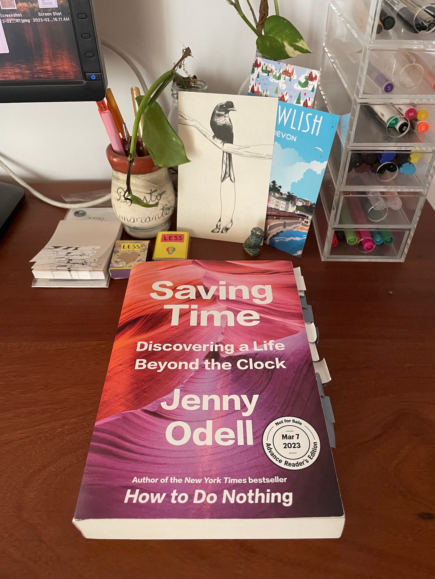 among stationery, poscards and plants, sits a book with post its stuck throughout. the title reads: saving time: discovering a life beyond the clock. jenny odell. author of the new york times bestseller how to do nothing. advance reasers edition, mar 7 2023