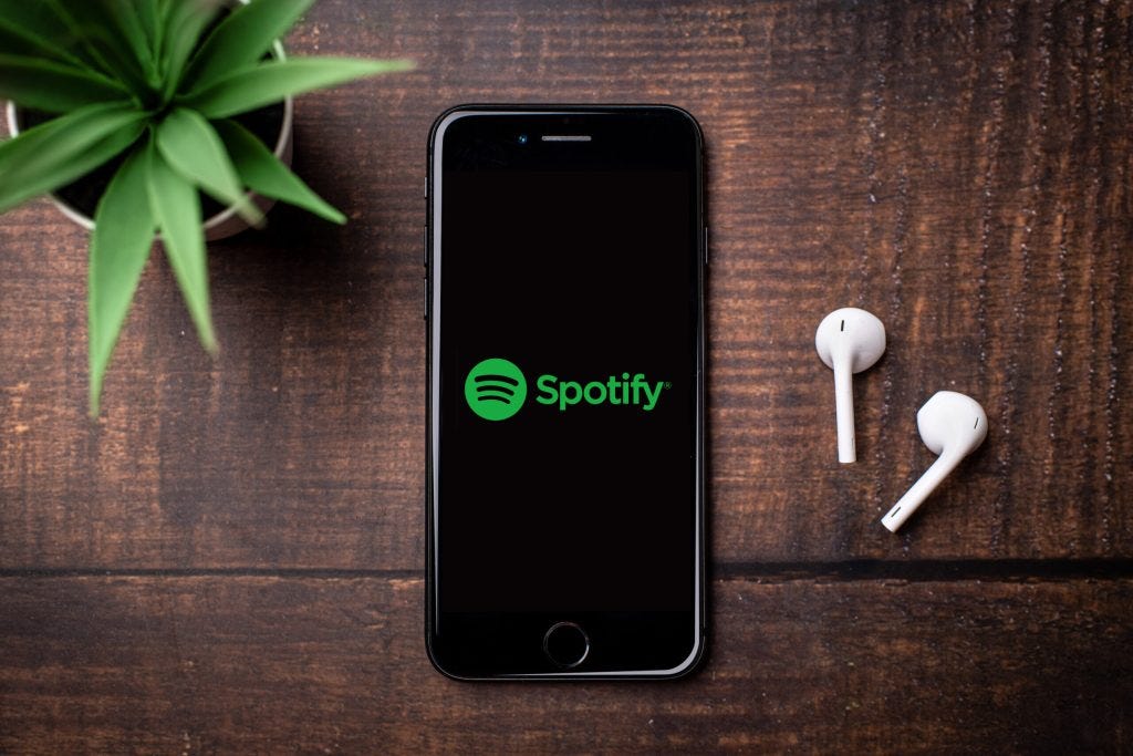A black iPhone displaying the Spotify logo on a wood table. To the left is a green succulent and to the right are Airpods.