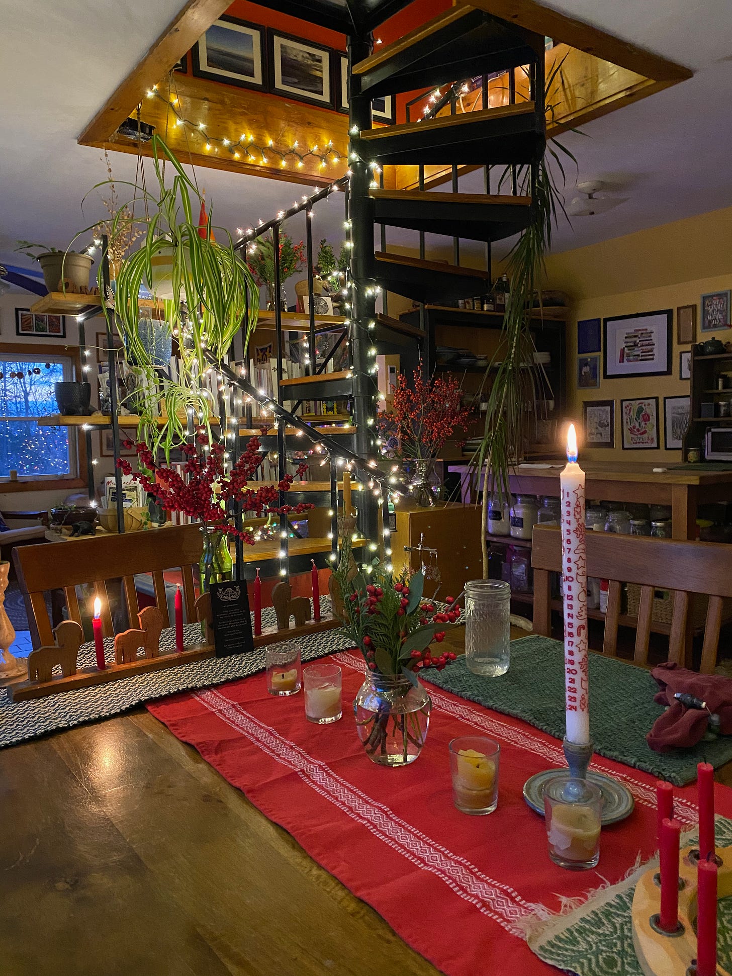 A table decorated with a red runner, jars of winterberry and greenery, and a tall white advent calendar. Behind the table, a spiral staircase strewn with twinkly lights.