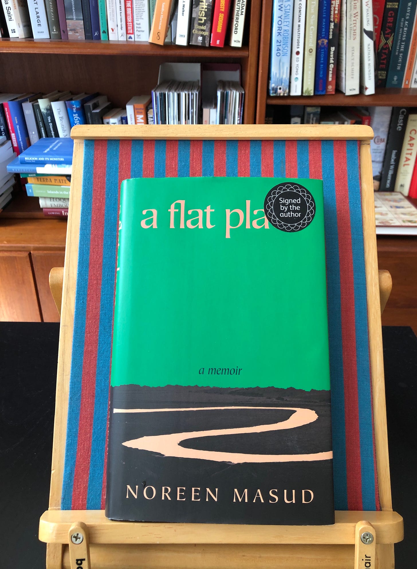 A book titled a flat place: a memoir, by Noreen Masud, sits on a stripy bookchair. A circular “signed by the author” obscures part of the title at the top right-hand corner. The cover is mostly blank, with a flat landscape along the bottom third, through which snakes an empty road. In the background is a wooden bookcase full of books.
