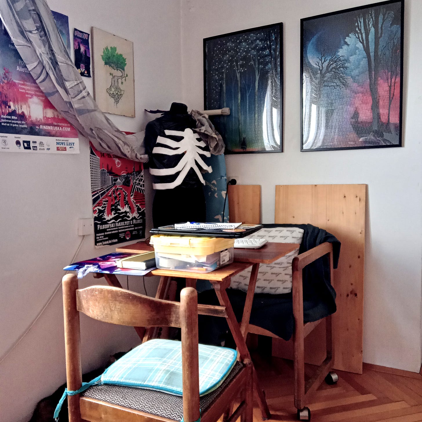 A corner of the living room with Rikon convention posters and Heye Puzzles by Andy Kehoe framed on the wall, a portable wooden balcony desk with a box of stationery which hold a closed laptop, two chairs, and a paper mache human ribcage on a black-cloth mannequin at the back, in the very corner.