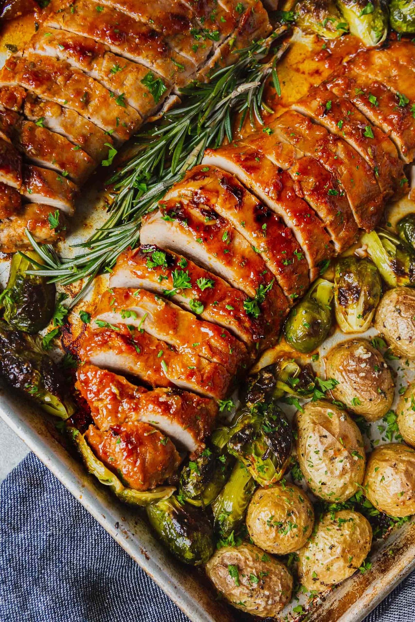 A sheet pan with two glazed, roasted pork loins. There are potatoes and brussels sprouts on the sheet pan also. The pork loins are sliced for serving.