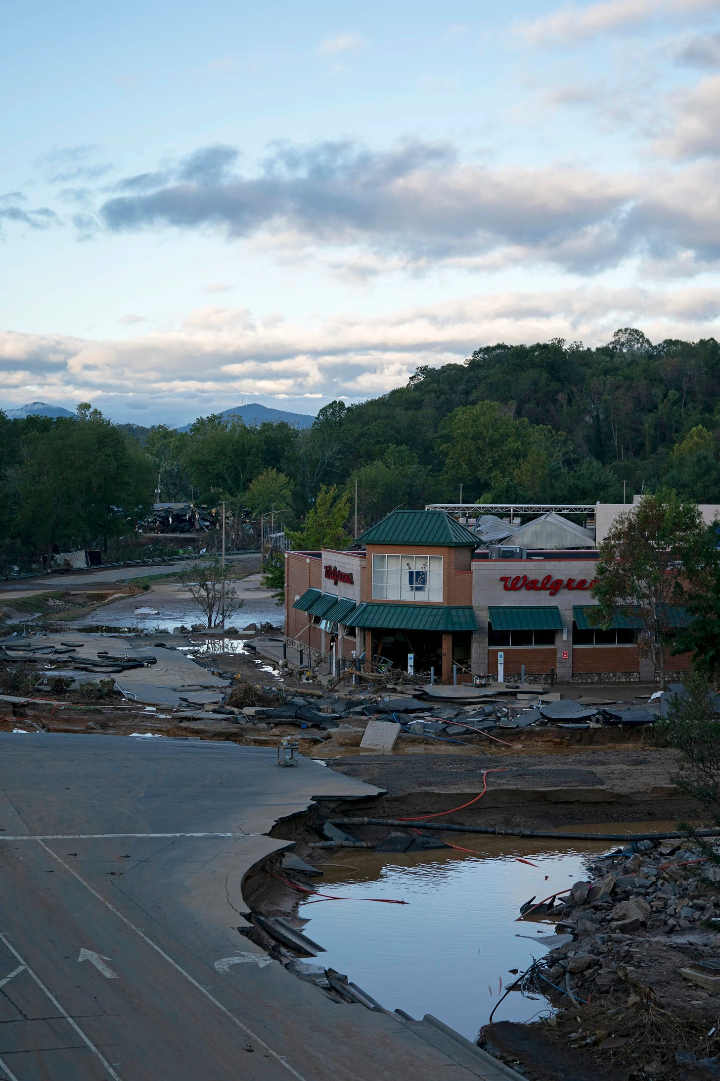 A flood-damaged Walgreens and surrounding area with debris, broken pavement, and water pools. Trees and hills are visible in the background