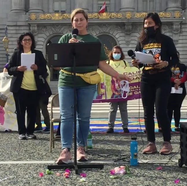 White women, Gretchen Sandler, speaking at a mic in front of SF City Hall with other women behind her. White women, Gretchen Sandler, speaking at a mic in front of SF City Hall with other women behind her.