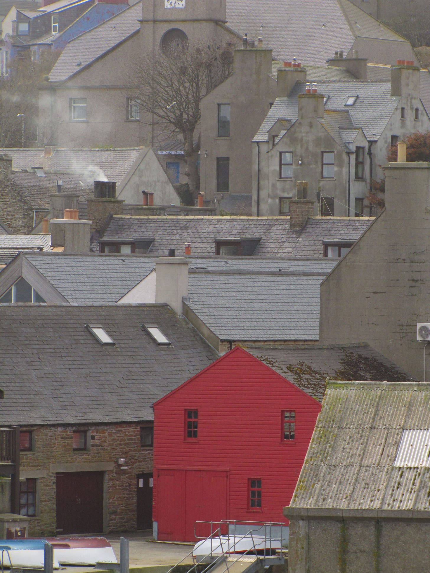 Houses and buildings climb up the hill of Brinkies Brae in Stromness, Orkney. One is painted red, but most are grey or beige, blending into the rock of the island. Smoke rises from one chimney and boats are overturned on a pier.