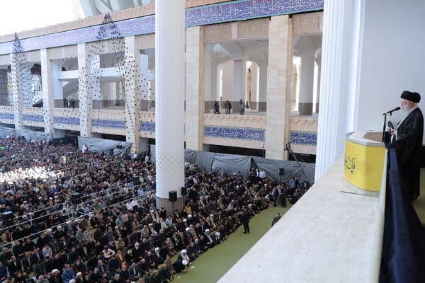 Ayatollah Ali Khamenei, standing behind a lectern on a podium, speaks to a large crowd of supporters.