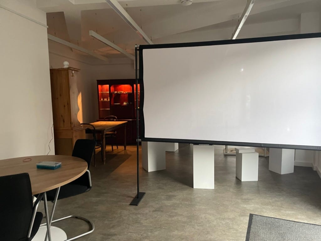 A small room with white walls. In the foreground there is a projector screen. To the left there is a dining room scene, lit warmly. 