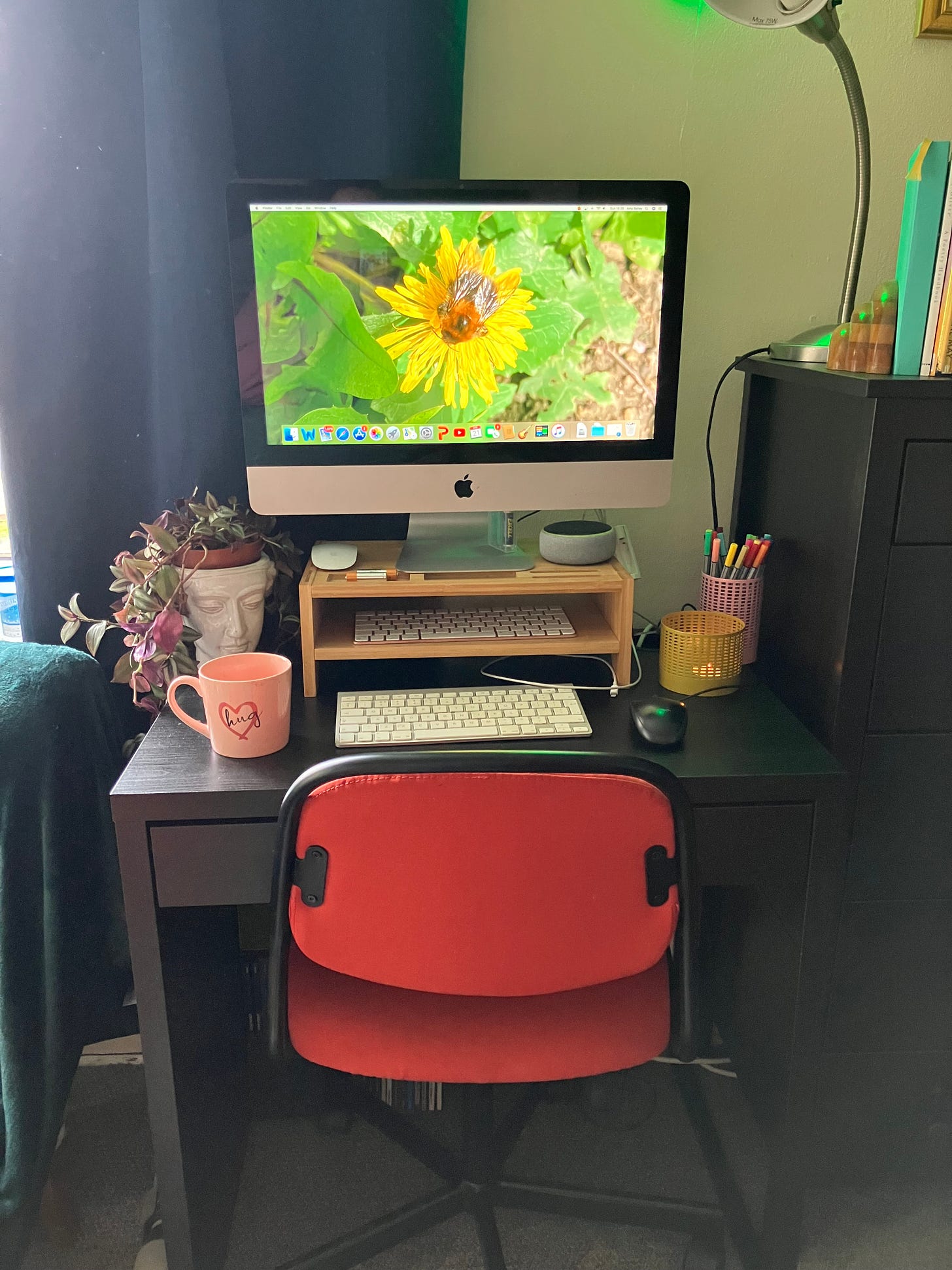 A very old iMac on a black desk, surrounded by a plant in a pot shaped like a face, a pink mug, a yellow tealight holder and a pink pen holder containing pens.  The desk chair is red.