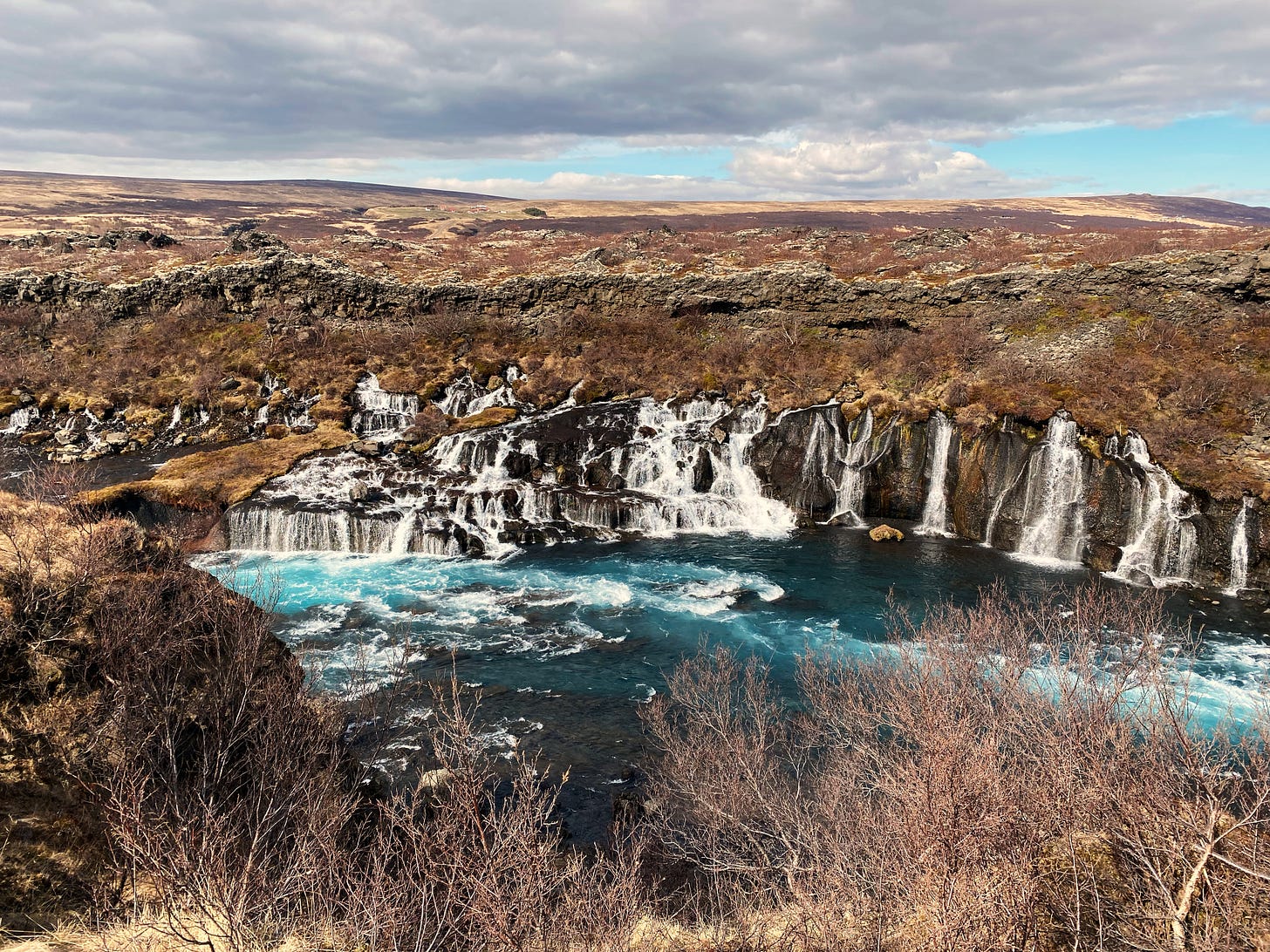 A blue sky with clouds stretches above a brown moss covered landscape.  In the foreground the Hraunfossar waterfall flows from lava rock directly into the blue river like a series of cascading bridal veils. 