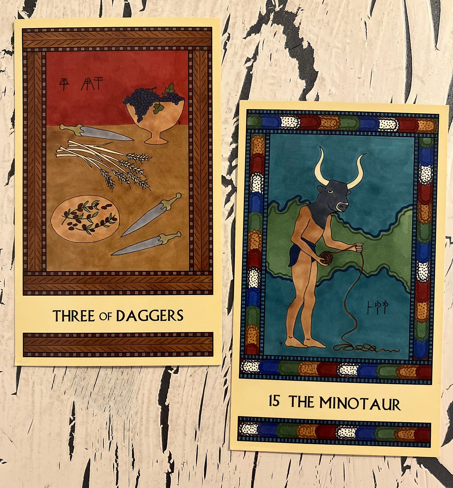 Two Minoan Tarot cards side by side on a crackled white background. The Three of Daggers is in shades of tan and deep red. It shows a tabletop with a bowl of grapes, a platter of olives, several stalks of wheat, and three daggers. The Minotaur has a multicolored border. It shows a Minoan man in a loincloth, wearing a bull's head mask and holding a ball of string that he's unwinding.