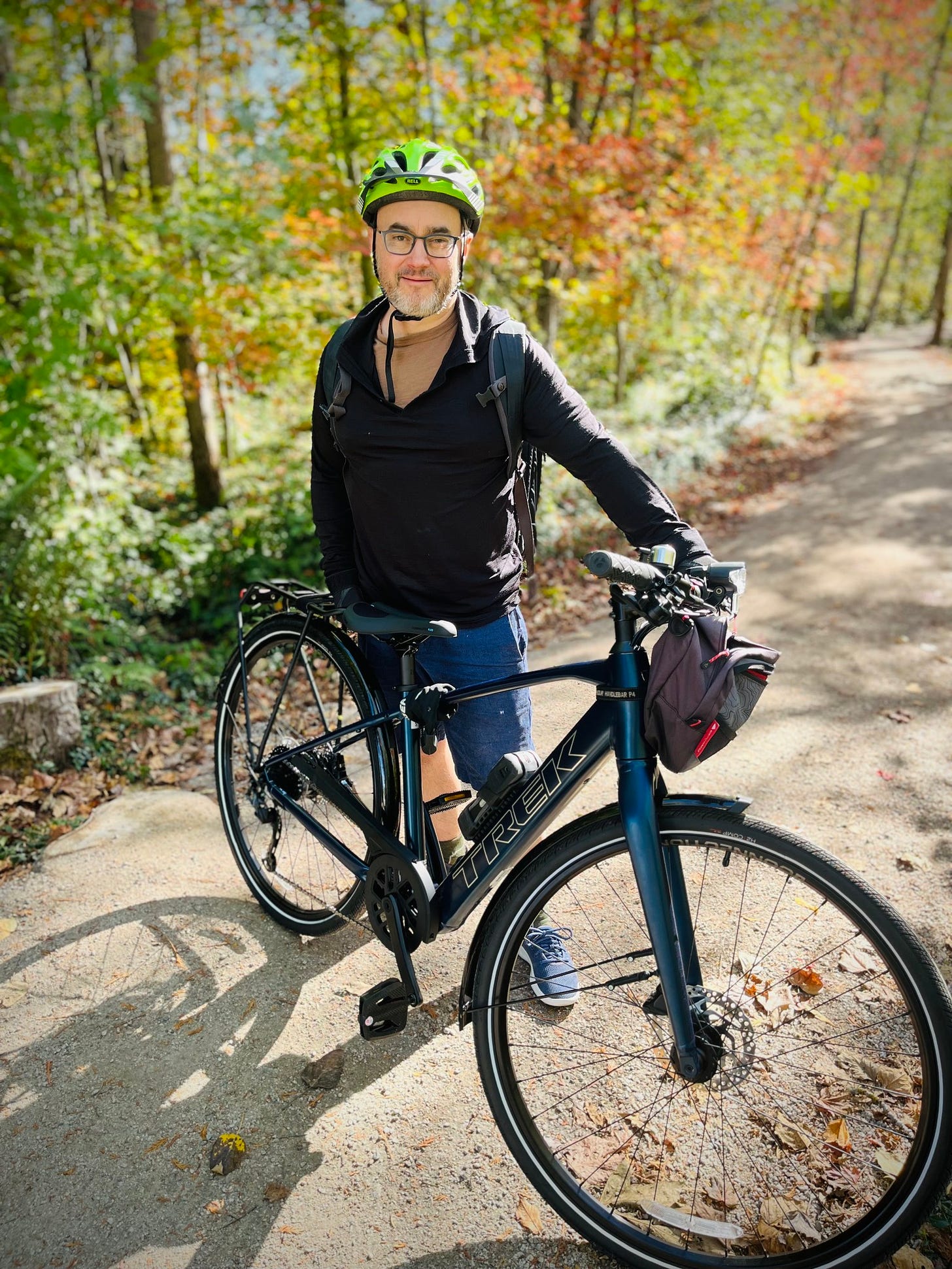 Photo of me standing behind a shiny new blue e-bike, on a forest path, with  very bright, sunlit, leafy forest behind me, with lots of autumn colour.