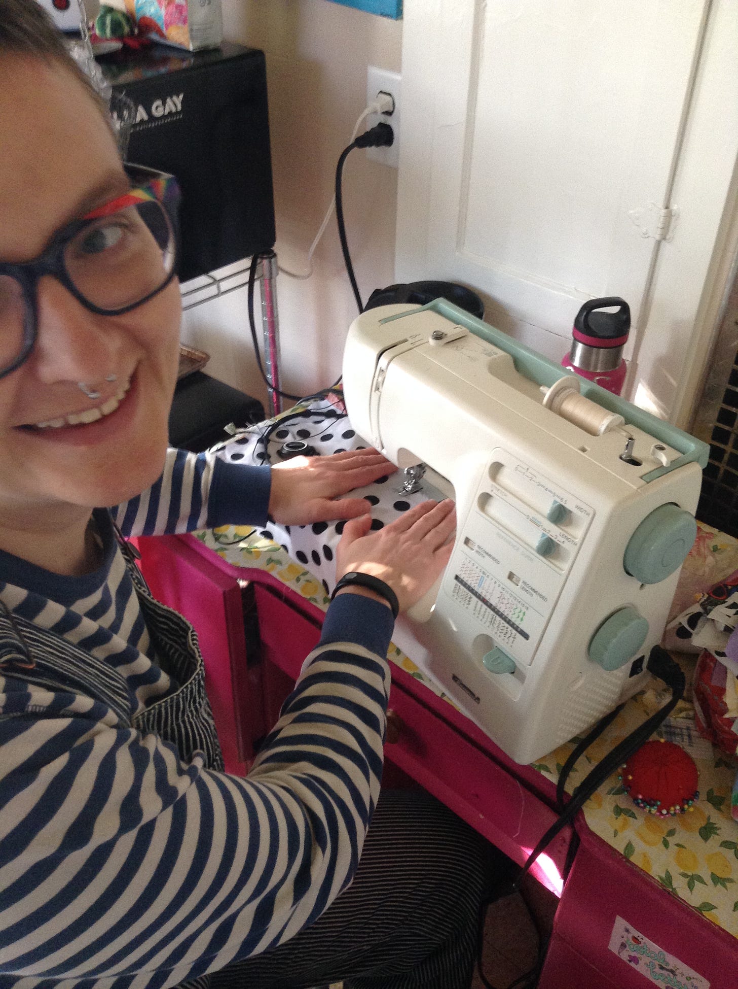 IMG DESCRIPTION: A white person sitting at a sewing machine, sewing a piece of black and white polka-dotted fabric. She is wearing a striped shirt and smiling widely.