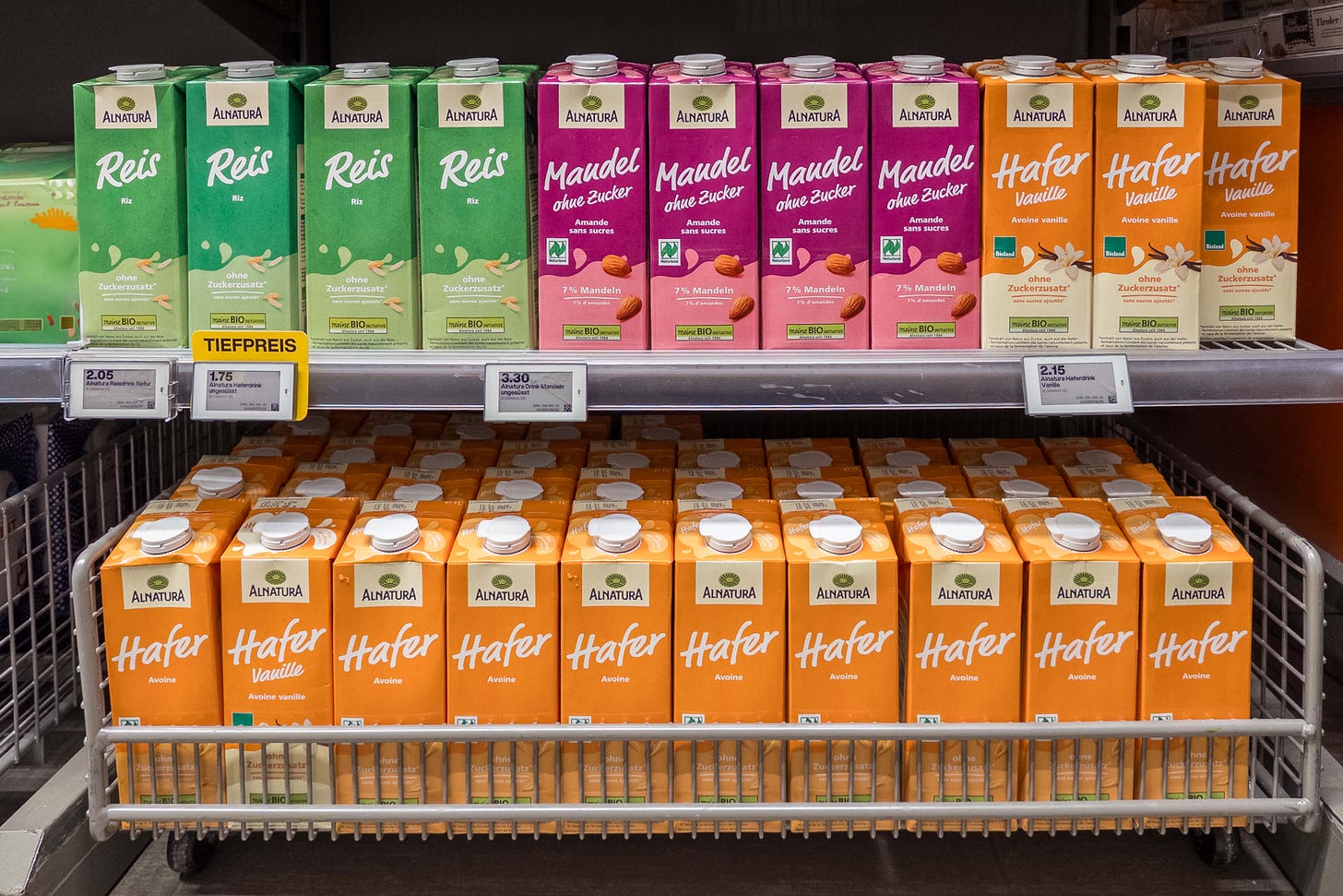 Supermarktregal mit dicht aneinander gereihten Getränkekartons der Marke Alnatura: Oben links grüne Packungen Reis-Drink, daneben pinkfarbene Mandel ohne Zucker-Drinks, rechts creme-orangefarbene Hafer Vanille-Drinks; eine digitale Preisanzeige zeigt Tiefpreise ab 1,75 CHF, darunter steht ein ganzer Wagen voller orangefarbener Hafer-Drinks. Supermarktregal mit dicht aneinander gereihten Getränkekartons der Marke Alnatura: Oben links grüne Packungen Reis-Drink, daneben pinkfarbene Mandel ohne Zucker-Drinks, rechts creme-orangefarbene Hafer Vanille-Drinks; eine digitale Preisanzeige zeigt Tiefpreise ab 1,75 CHF, darunter steht ein ganzer Wagen voller orangefarbener Hafer-Drinks.