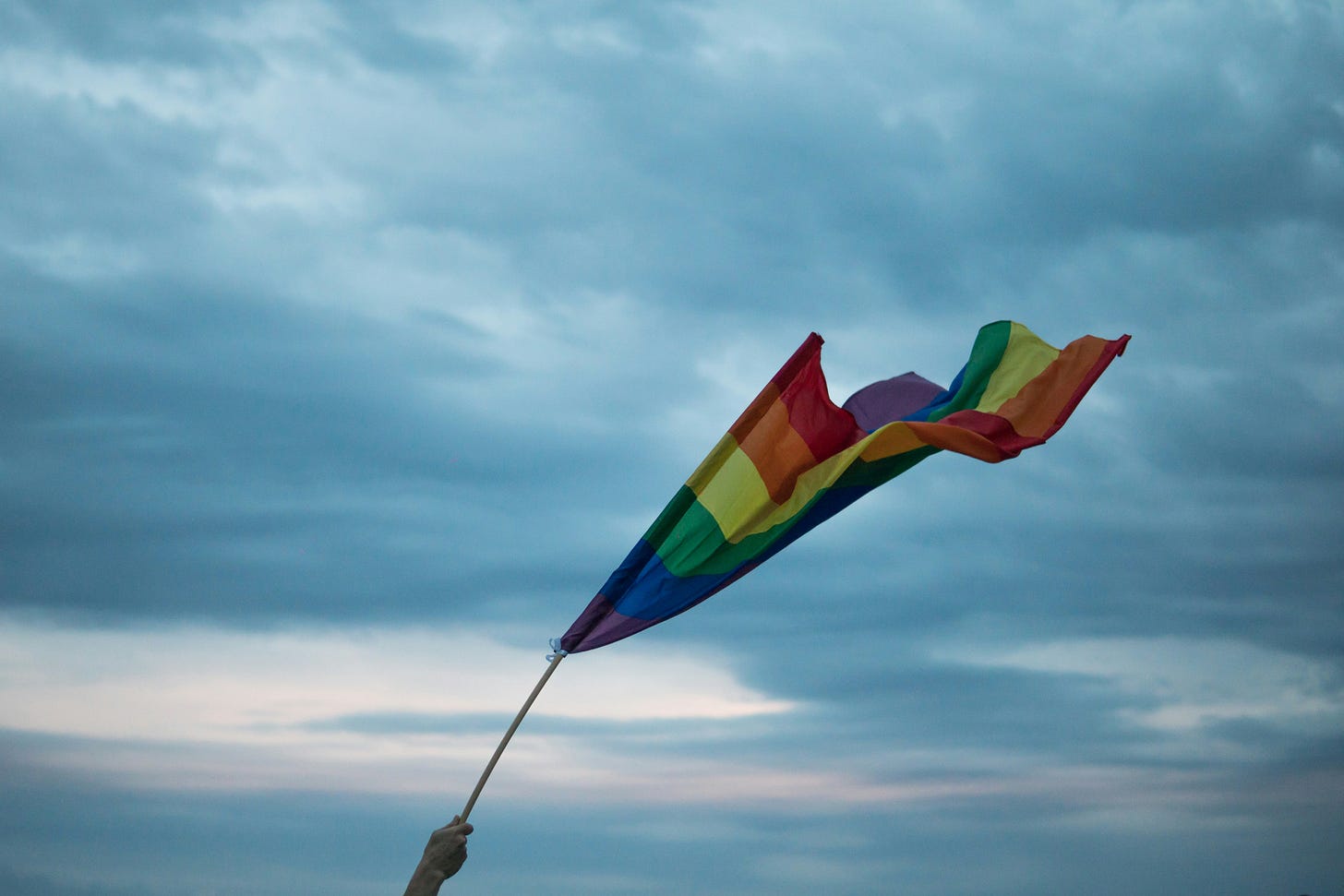 against a backdrop of gloomy clouds, a hand clutches to a rainbow flag, straining against the wind