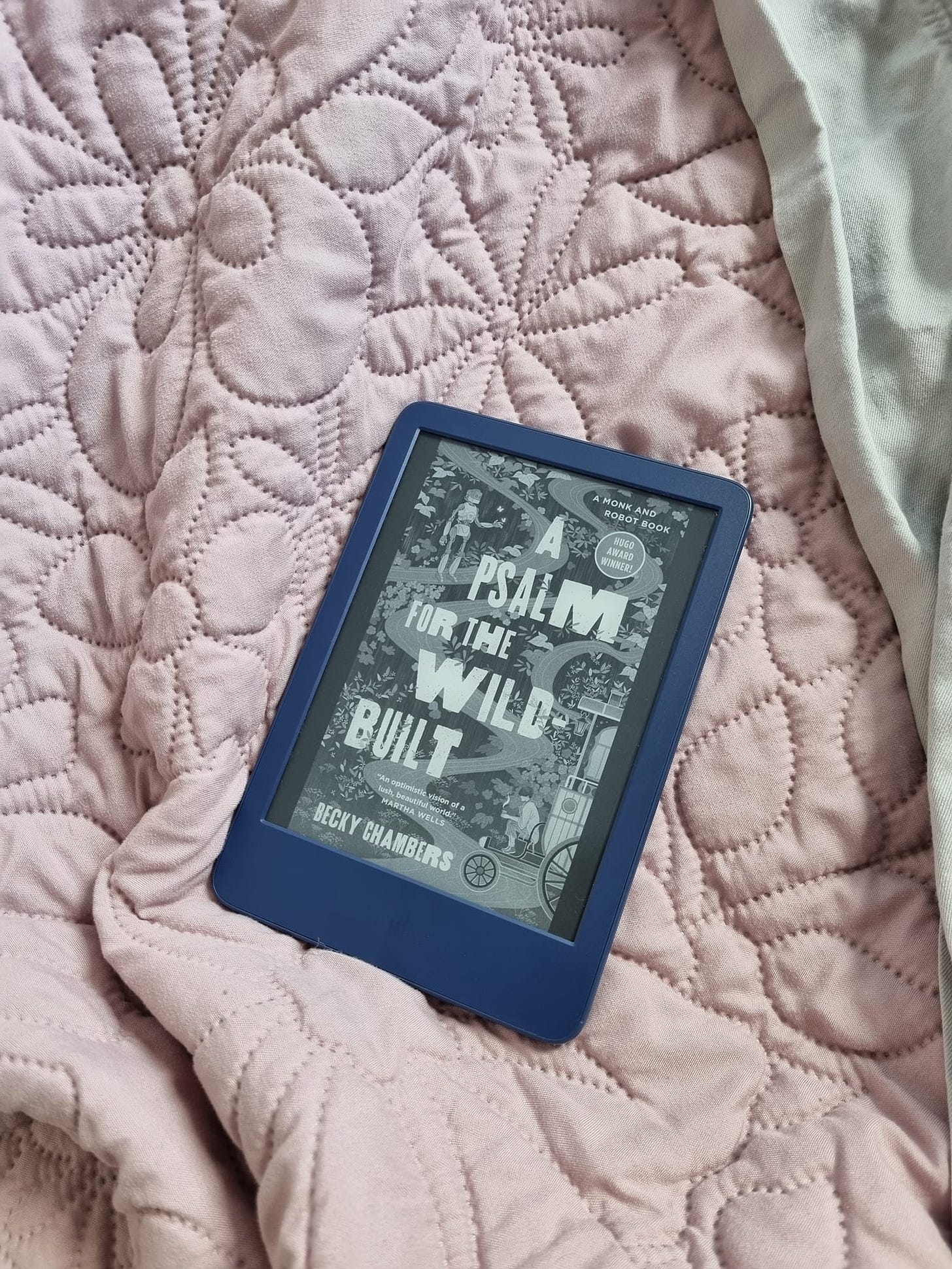A Kindle lying on a pink bedspread showing the cover of A Psalm for the Wild-Built by Becky Chambers