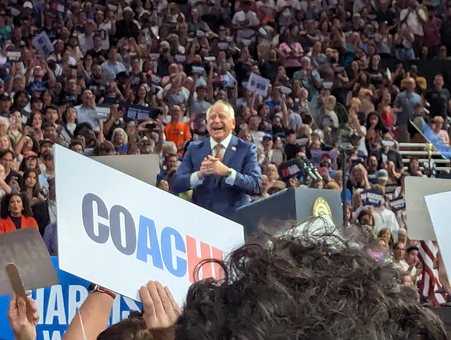 Tim Walz smiling and clapping on stage in a totally packed arena