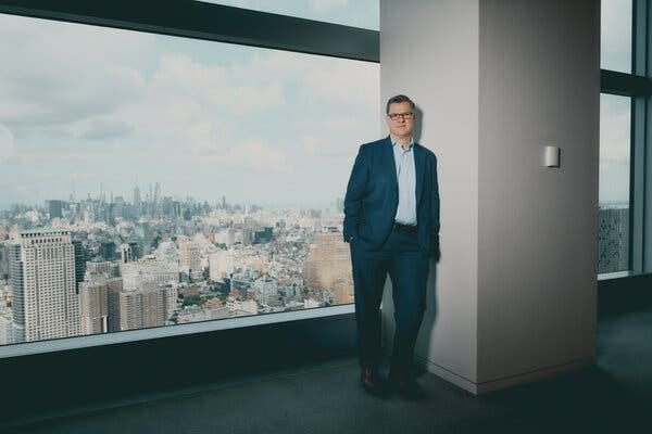 Jim Covello, wearing a blue suit, stands near a big window with a view of the New York City skyline.