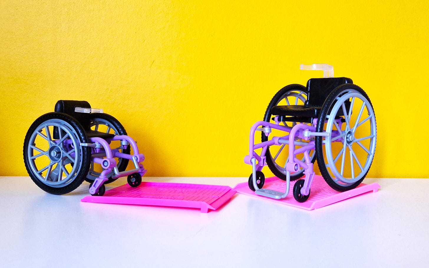 Two Barbie wheelchairs, both with purple frames and each of them with their individual hot pink colored ramp, staged before a bright yellow wall on a white table.