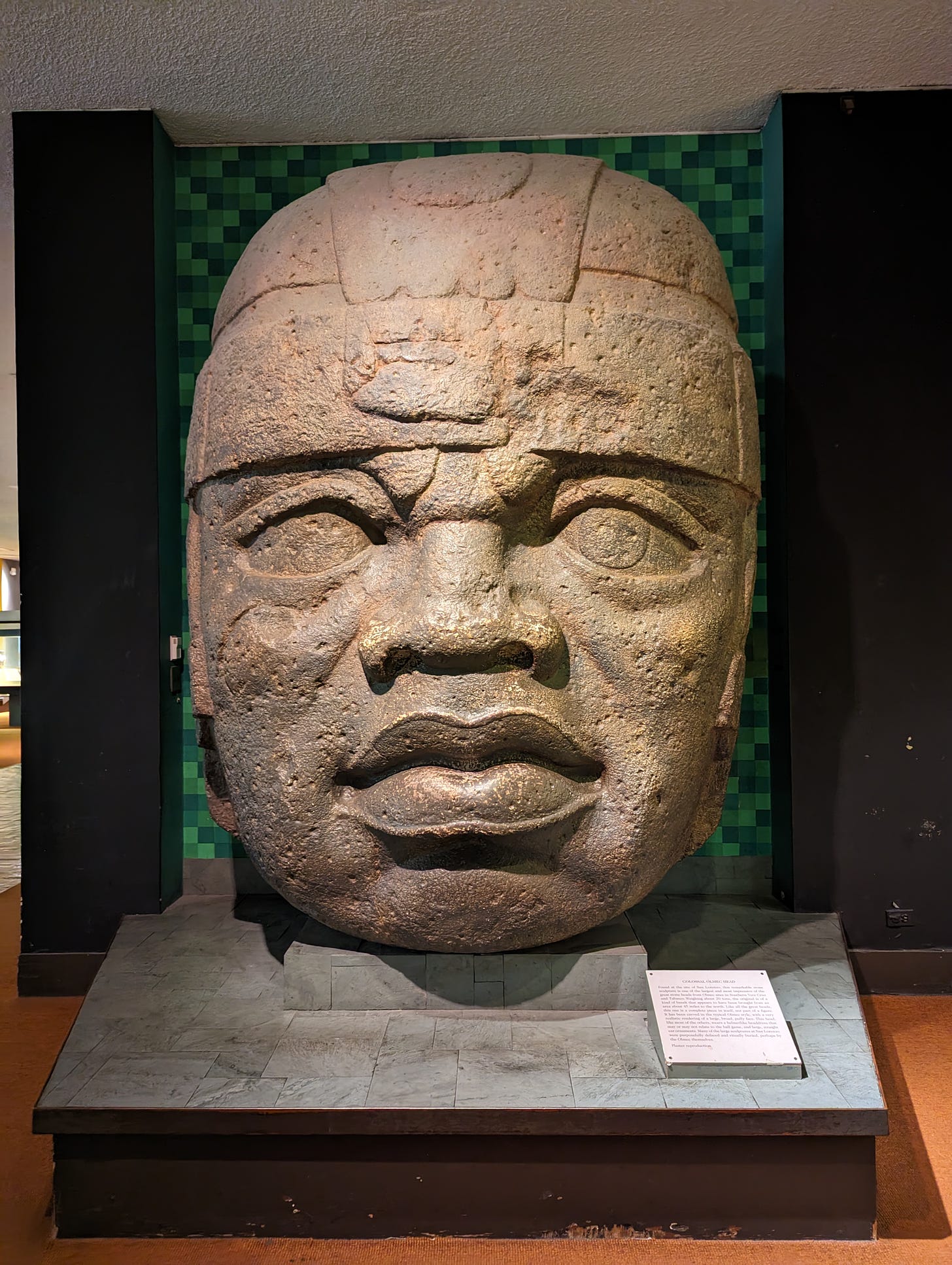 Plaster reproduction of a colossal Olmec head from San Lorenzo, Mexico. The head bears the hallmarks of traditional Olmec carving: a broad, puffy face, realistic features, a helmet or headdress, and ear decorations.