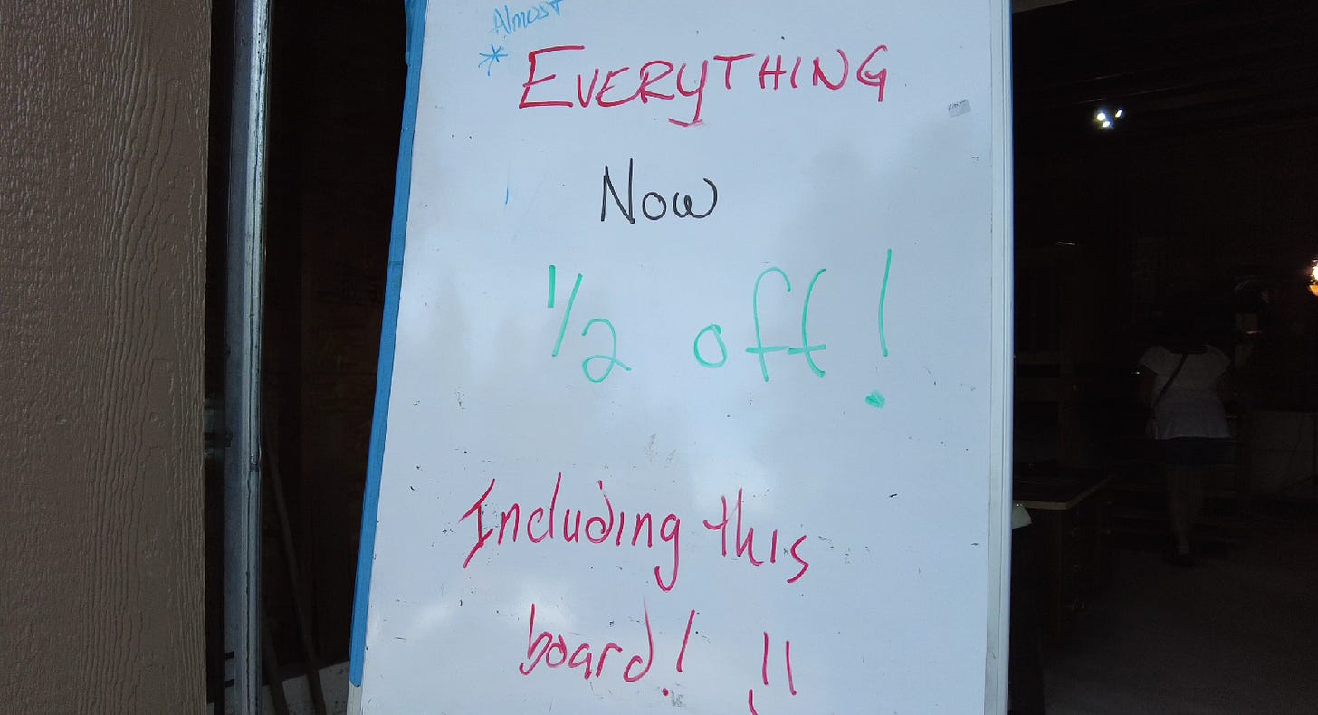 white board sign at a yard sale: "Everything now 1/2 off, including this board!!"