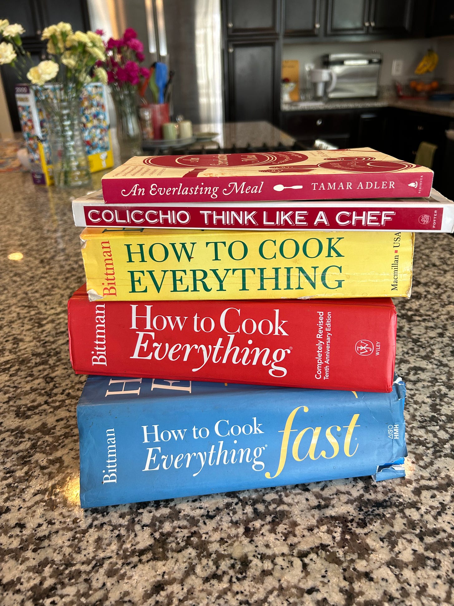 Four cookbooks are stacked on a kitchen counter. From bottom to top they are How To Cook Everything Fast, one red and one yellow How to Cook Everything, Think Like a Chef, and An Everlasting Meal