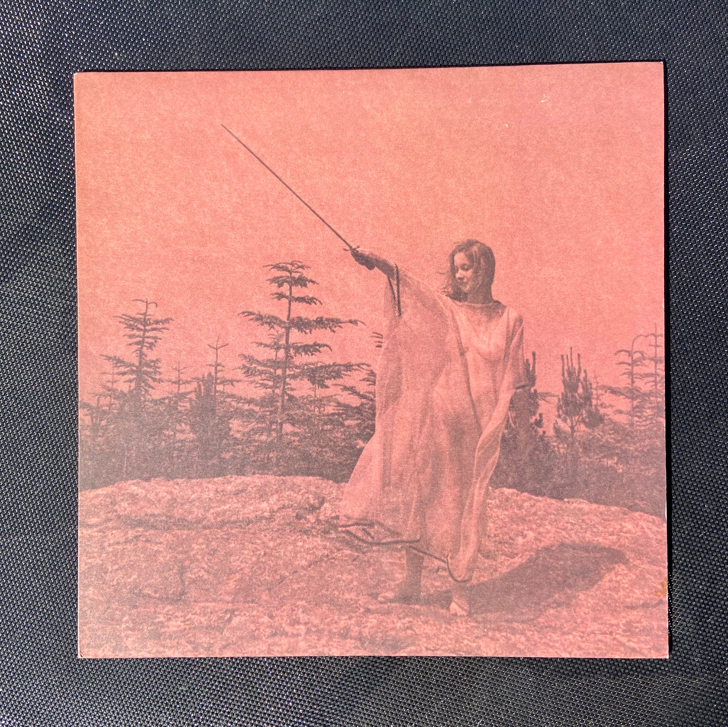 the front album cover of Unknown Mortal Orchestra's II. a pale pink album with an image of a woman in a sheer dress holding a sword on a mountaintop in front of trees