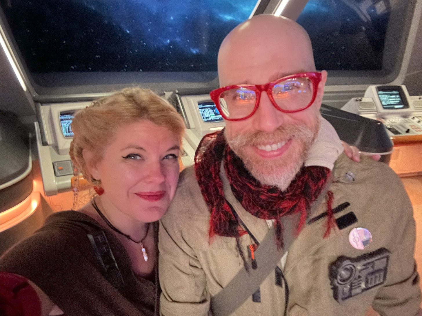 Blonde woman and bald man with red glasses, both dressed in shades of red and brown, standing in front of a bunch of starship consoles with a starfield visible through the windows