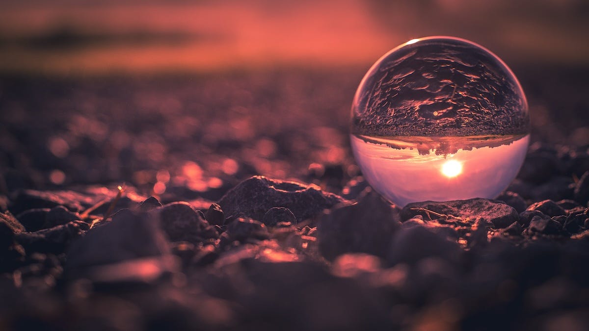 A small crystal ball sits on the ground and reflects an upside down image of the sky, sun and landscape.