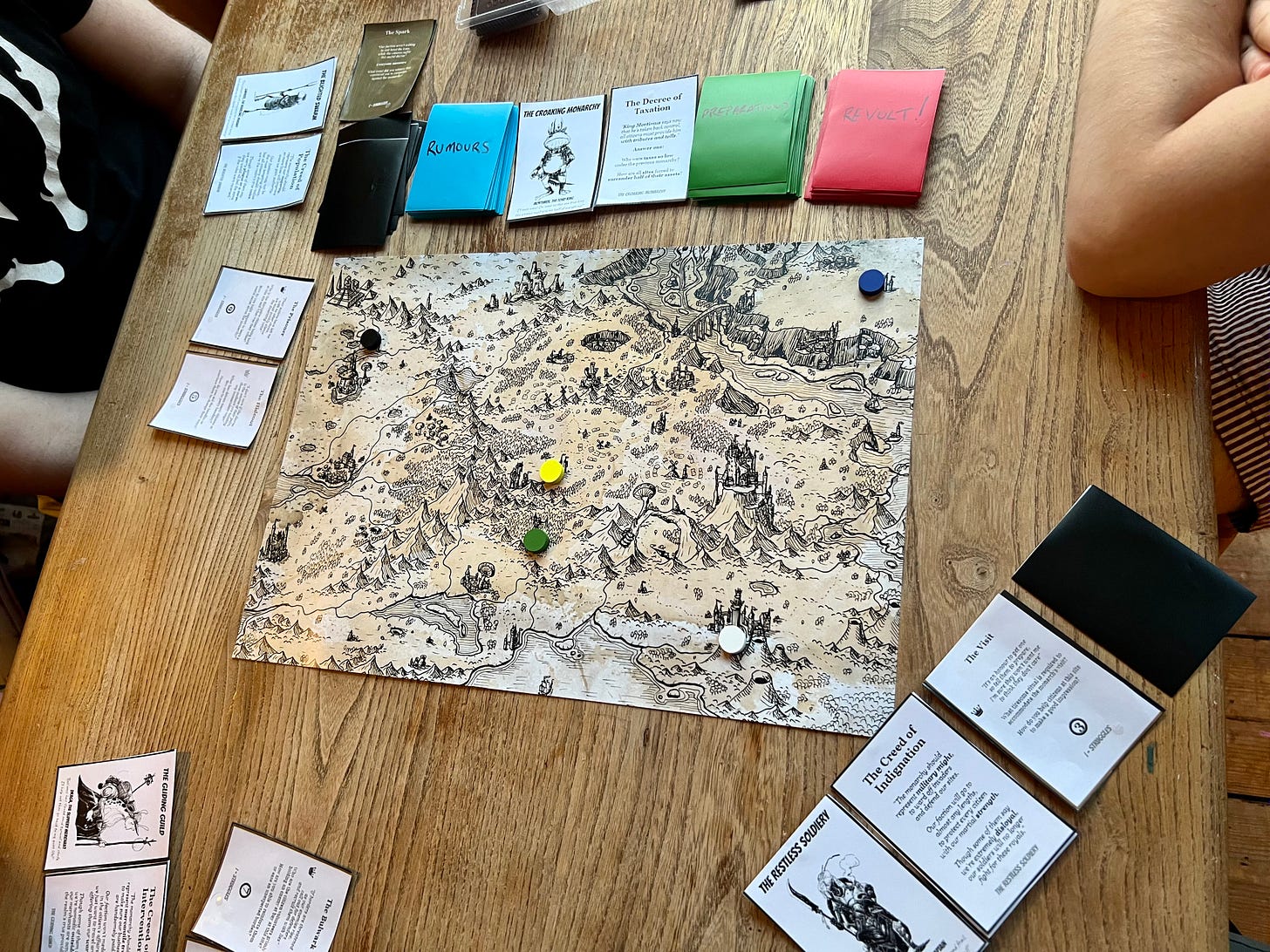 A black and white map of a kingdom, surrounded by playing cards with players sat around the table,
