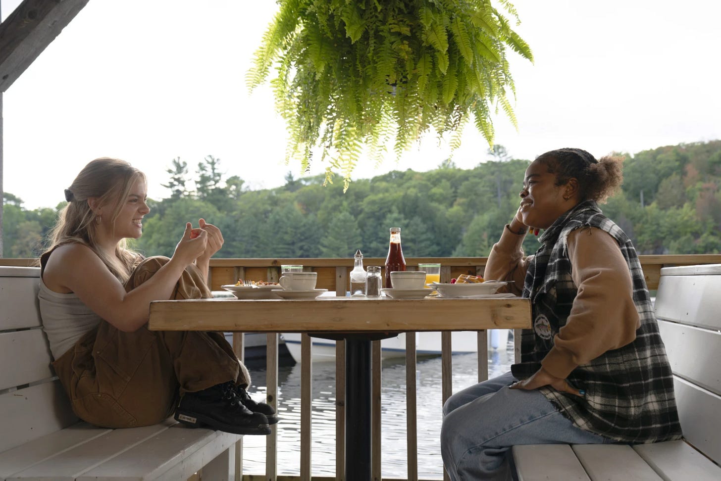 Two women sit at a table having breakfast. There's a lake and marina behind them.