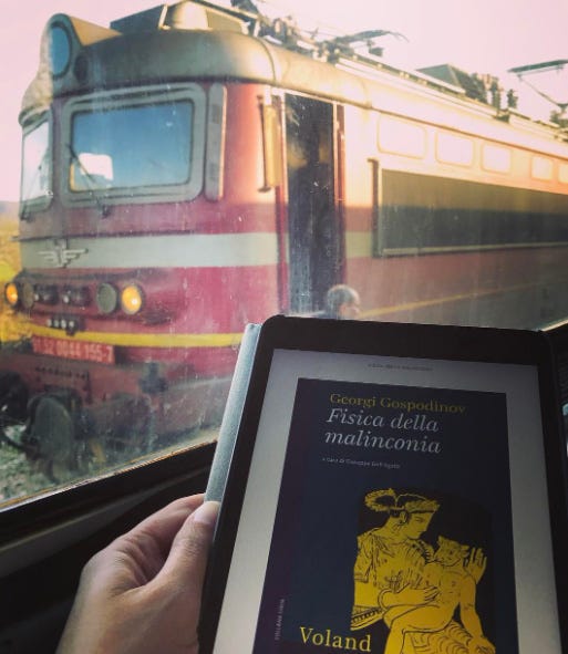 copertina di un libro digitale in primo piano e un vecchio treno bulgaro di colore rosso visto attraverso il finestrino di un altro treno