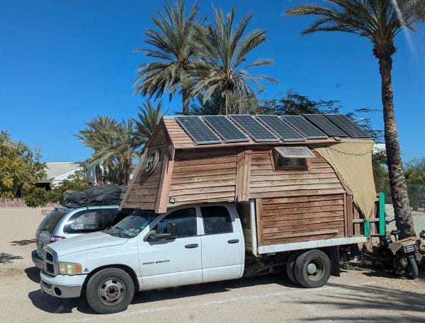 a white pickup truck, heavily burdened and riding low under the weight of a homemade wooden house with solar panels on the roof