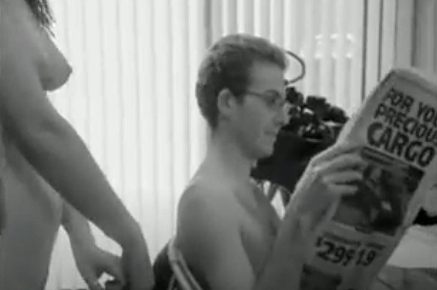 A nude man sits reading a newspaper. Just barely out of frame on the left a nude woman appears to be standing over him. 