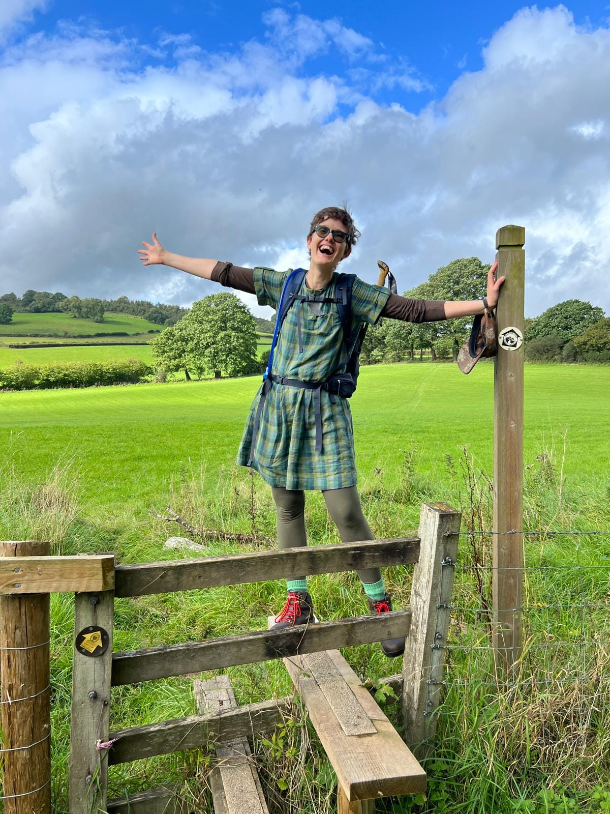 Katie, a white human stands on a stile at the entrance to a vibrant green field. Katie’s arms are outstretched in a gesture of expansive delight.