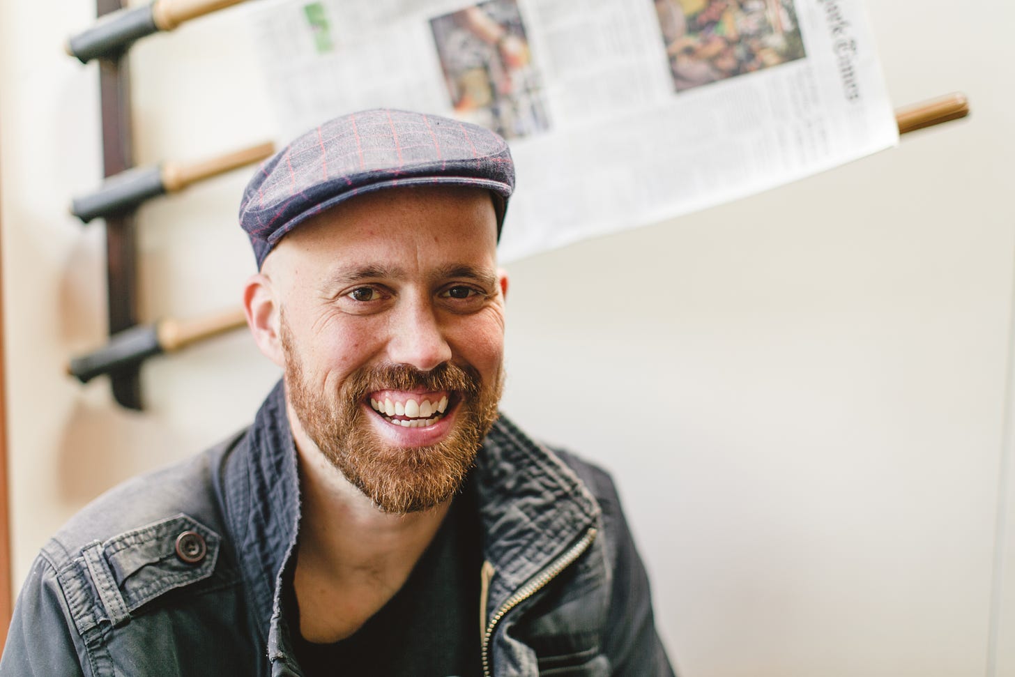 A photo of a white man with a reddish beard, wearing a newsboy cap against the background of a white wall