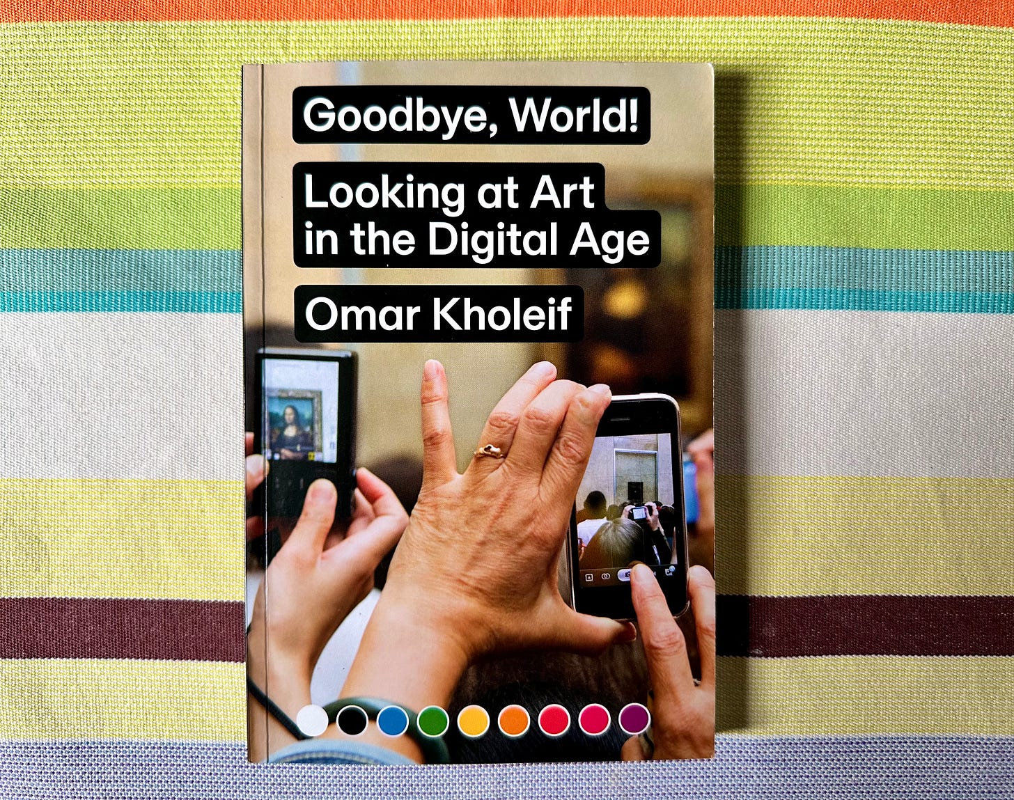 Image of the book "Goodbye, World! Looking at Art in the Digital Age" by Omar Kholief on a green, turquoise and yellow multi striped cloth. The cover of the book shows the hands of a man in the foreground taking a photon a crowd which on close examination appears to be the Mona Lisa, blurred in the background, surrounded by lots of other people out of focus doing the same. White text is overlaid stating “First #3: Goodbye, World! Looking at Art in the Digital Age. Digital culture, art, and how technology shapes our perception of each.” Available February 1st, 2024 at UXMICHAELCO.SUBSTACK.COM”.